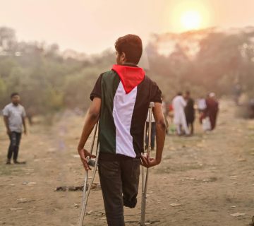 A young man wrapped in a Palestinian flag walks with crutches through a field in Bangladesh during the day.
