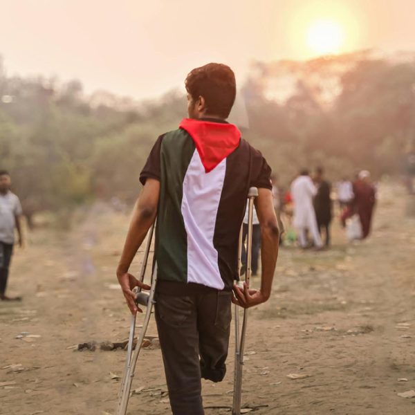 A young man wrapped in a Palestinian flag walks with crutches through a field in Bangladesh during the day.